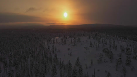 Aerial-view-of-a-hazy-sun-behind-silhouette-hills-and-snowy-forest-of-Lapland