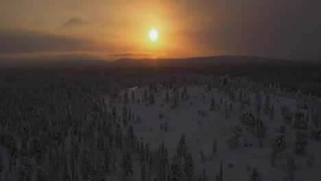 Vista-Aérea-De-Un-Sol-Brumoso-Detrás-De-Colinas-De-Silueta-Y-Bosques-Nevados-De-Laponia