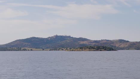 A-serene-ground-level-view-of-Monsaraz-as-seen-from-the-lakeside,-highlighting-the-tranquil-waters-and-the-picturesque-medieval-village-perched-atop-the-hill-in-the-background