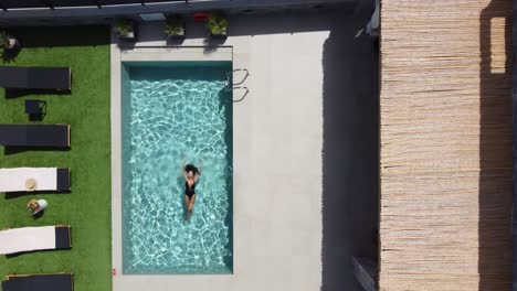 Drone-flies-over-an-outdoor-pool-in-bird's-eye-view---Woman-in-swimming-costume-floats-on-her-back-on-water---Villa-in-Greece-Crete-with-palm-trees