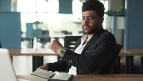 portrait-of-a-handsome,-stylish-young-man-of-Arab-descent-sitting-in-a-modern-business-center-office-with-a-laptop,-putting-on-glasses-for-vision-correction