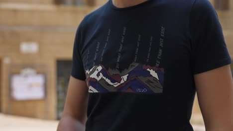 A-man-is-shown-wearing-a-t-shirt-featuring-a-mountain-scene,-with-a-small-device-clipped-onto-it,-possibly-for-audio-or-recording-purposes,-captured-outdoors-in-urban-setting