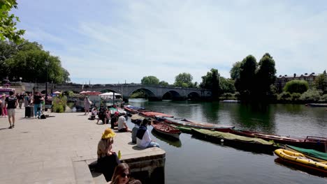 Richmond,-London:-View-of-people-walking-along-the-Thames-riverbanks-with-the-bridge-in-the-background,-enjoying-the-scenic-surroundings-and-pleasant-weather