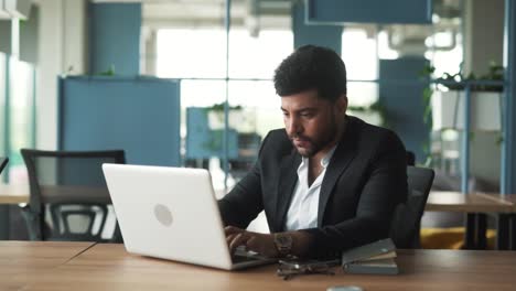 portrait-of-a-handsome,-stylish-young-man-of-Arab-descent-sitting-in-a-modern-office-of-a-business-center,-working-intently-on-his-laptop-and-contemplating