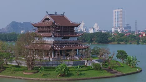 Este-Metraje-De-Un-Dron-Captura-Un-Templo-Tradicional-Situado-En-Una-Pequeña-Isla,-Con-El-Telón-De-Fondo-De-Una-Ciudad-Moderna-En-Ninh-Binh,-Vietnam.