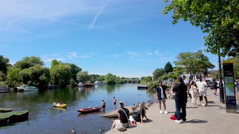 Richmond,-London:-View-of-people-sunbathing-on-the-Thames-riverbanks-with-the-bridge-in-the-background,-enjoying-the-sunny-weather-and-scenic-surroundings