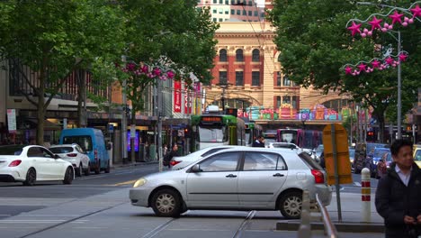 Vehicle-traffic,-pedestrians-crossing-and-trams-running-on-Elizabeth-street-with-tree-lined-street-decorated-with-festive-decorations-in-the-bustling-city-of-Melbourne,-a-vibrant-urban-street-scene