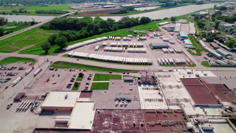 Aerial-view-showing-semi-trucks-on-the-road-and-a-large-parking-lot-filled-with-trailers,-alongside-industrial-buildings-and-greenery