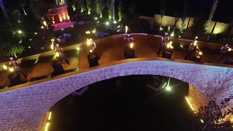 Aerial-clip-of-actors-playing-drum-using-torch-near-the-illuminated-bridge-on-a-luxury-venue-for-a-wedding