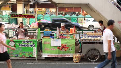 Bangkok,-Thailand---Thailändische-Verkäuferinnen-Verkaufen-Essen-Und-Getränke-Entlang-Der-Straße-An-Der-Pratunam-Brücke,-Im-Hintergrund-Fahren-Transportfahrzeuge-Vorbei---Nahaufnahme