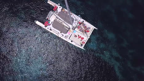 Top-down-drone-shot-of-a-catamaran-hovering-over-the-deep-ocean-on-crystal-lagoon-at-comino-island-in-malta-with-many-people-enjoying-themselfs