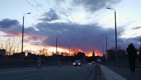 Los-Autos-Pasan-Por-El-Puente-Deglava-Con-Una-Hermosa-Puesta-De-Sol-Naranja-Y-Una-Silueta-De-Iglesia-En-El-Fondo,-Letonia