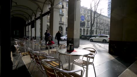 People-passing-by-on-arch-covered-pavement,-empty-tables-and-chairs