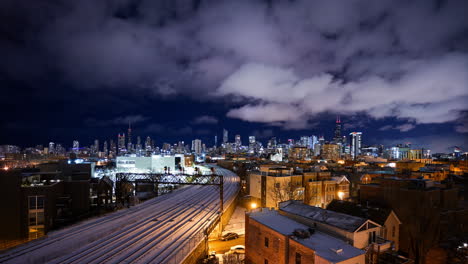 Night-scene-with-white-puffy-clouds-passing-over-a-snow-covered-Chicago-skyline-with-trains-rolling-by-on-the-tracks-time-lapse