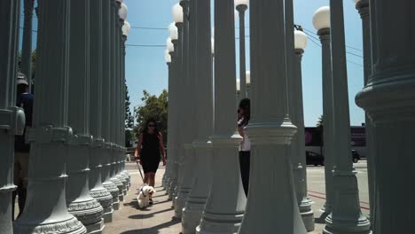 slow-motion-shot-of-female-walking-towards-camera-with-dog,-at-LACMA-lights,-during-the-sunny-day,-Los-Angeles