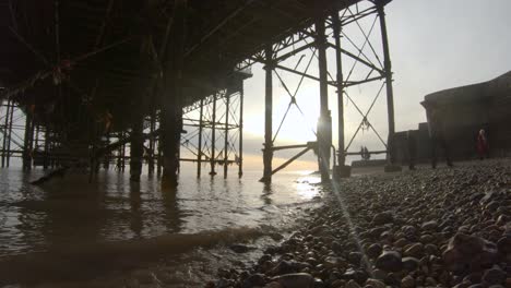 Sun-setting-and-waves-splashing-under-Brighton-pier-in-the-UK-with-sun-flare-and-people,-slow-mo