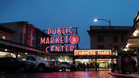 Neonreflexion-Von-Regnerischen-Pfützen-Auf-Der-Straße-Vor-Dem-öffentlichen-Marktzentrum-Und-Der-Pike-Place-Fish-Company---Dunkler,-Launischer-Himmel-Im-Hintergrund,-Vögel-Und-Straßenlaternen