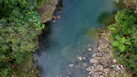 People-relaxing-on-river-bank-of-Angelito-Trail-rainforest,-overhead-rising-aerial