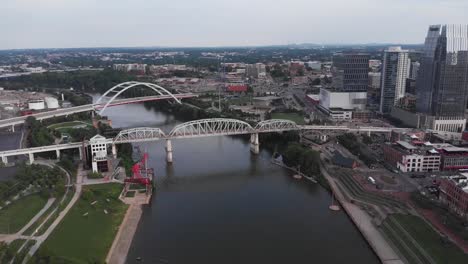 Truss-bridges-above-narrow-river-by-downtown-Nashville-city,-aerial-approach