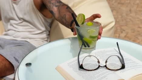 Man-on-holiday-relaxing,-enjoying-a-Caipirinha-cocktail-on-bean-bag-cushion-reading-a-book