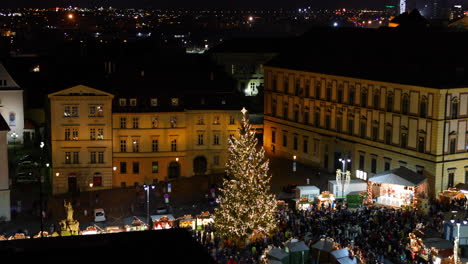 Luftaufnahme-Des-Dominanten-Weihnachtsbaums-In-Brünn-Auf-Einem-Der-Plätze,-Die-Die-Atmosphäre-Der-Märkte-Und-Einen-Stand-Mit-Vielen-Besuchern-Der-Veranstaltung-Zeigt,-Aufgenommen-In-4K-60fps-Zeitlupe