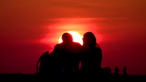 A-couple-playfully-sitting-at-North-Avenue-Beach-in-Chicago-kissing-in-front-of-a-bright-yellow-sun-with-orange-sky