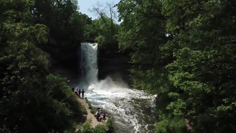 Wide-aerial-shot-in-slow-motion-slightly-rotating-while-looking-up-at-the-Minnehaha-waterfall-surrounded-by-trees-with-a-family-of-tourists-looking-at-the-falls-from-a-trail-below