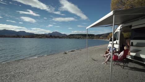 SLOWMO---Young-couple-sitting-outside-at-campervan-on-beach-by-Lake-Wanaka,-New-Zealand