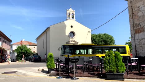 Un-Autobús-Urbano-Amarillo-Se-Encuentra-Frente-A-La-Iglesia,-Esperando-A-Los-Transeúntes-En-Prementura-Croacia