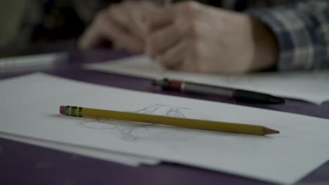 Sketch-pencils-sitting-on-top-of-purple-table-while-sketch-artist-in-checkered-shirt-draws-on-sketch-paper-in-the-background