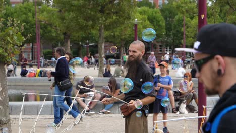 Slow-motion-footage-of-Man-in-black-top-and-child-in-green-t-shirt-create-bubbles-with-wand-in-park-while-child-looks-on,-Amsterdam,-Netherlands