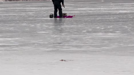 Una-Panorámica-Vertical-Desde-Un-Lago-Congelado-Hasta-Un-Hombre-Acercándose-A-Su-Equipo-De-Pesca-En-Hielo-En-Un-Lago-Congelado