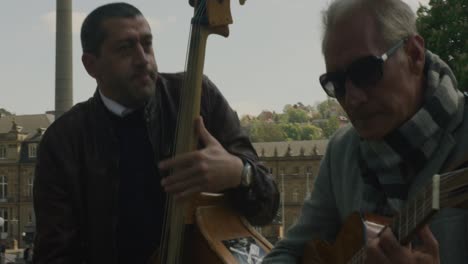 Tres-Músicos-Callejeros-Tocando-Instrumentos-Frente-A-La-Famosa-Estatua-Balvaria-En-La-Plaza-Del-Centro-De-Stuttgart-Al-Mediodía,-Alemania,-Europa,-ángulos-De-Visión-Panorámica