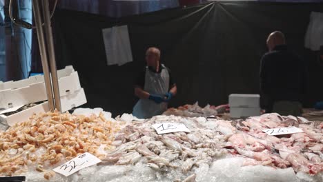 Slow-motion-close-up-on-shrimps,-squid-and-fish-on-ice-in-the-fresh-fish-market,-with-busy-people-working-behind-it