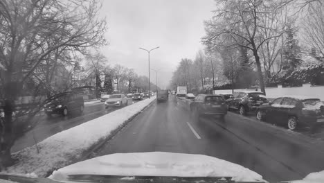 Driving-by-car-through-busy-Munich-city-at-a-wet-winter-day-with-driving-cars-in-front-of-the-drivers-view-through-the-snowflakes-droped-windshield-in-black-and-white