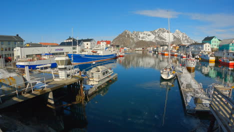 Mountains-reflecting-on-the-water-surface-in-Henningsvar-Harbor-on-a-sunny-calm-day,-Lofoten,-Norway,-panning-shot