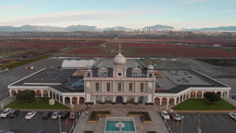 Imágenes-De-Drones-4k-Tomadas-Hacia-Adelante-Fachada-Frontal-Del-Templo-Sikh-Con-Bellas-Y-Elaboradas-Esculturas-Y-Arquitectura-Simétrica-En-Medio-De-Una-Tierra-De-Cultivo-Campos-De-Arándanos-Montañas-Cielo-Azul