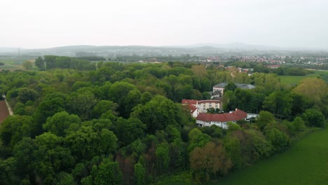 Aerial-view-towards-forest-and-a-villa,-a-town-in-background,-cloudy-day,-in-Germany---Approaching,-drone-shot