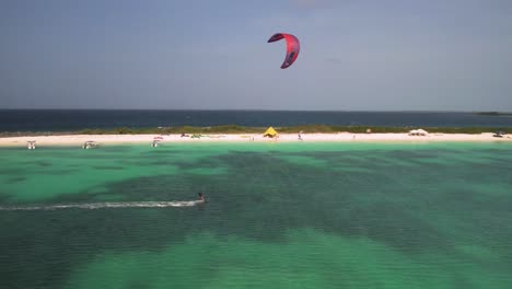 A-kite-surfer-gliding-over-crystal-clear-turquoise-waters-near-a-sandy-beach,-aerial-view