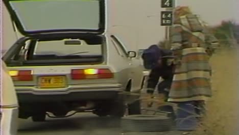 1970S-POLICE-OFFICER-HELPING-CHANGE-A-TIRE-ON-THE-SIDE-OF-THE-ROAD