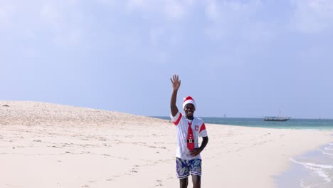 African-Man-Wearing-Santa-Hat-And-Tie,-Waves-At-Drone-Camera-While-Walking-At-Kwale-Sandbank-In-Tanzania