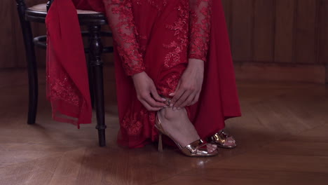 Woman-in-elegant-red-lace-dress-adjusting-her-gold-high-heels,-preparing-for-an-event