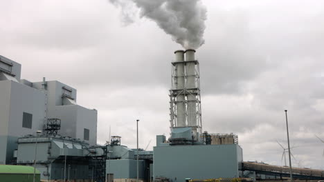 Smoke-rises-from-factory-smokestack-in-Eemshaven,-Netherlands,-slow-motion,-wind-turbines-spinning-in-background,-highlighting-contrast-between-dirty-and-clean-energy