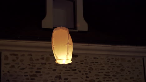 sky-lantern-glows-warmly-as-it-floats-in-front-of-a-rustic-building-at-night,-symbolizing-peace