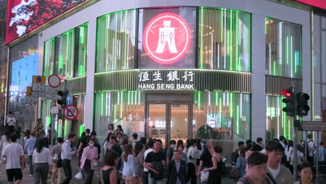 Una-Gran-Multitud-De-Peatones-Pasa-Junto-A-Una-Sucursal-Del-Hang-Seng-Bank,-Una-Empresa-De-Servicios-Bancarios-Y-Financieros-Con-Sede-En-Hong-Kong-E-Indicador-Del-Mercado-De-Valores-De-Hong-Kong,-Durante-La-Noche-En-Hong-Kong.