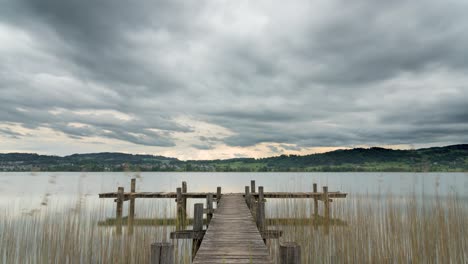 T-shaped-wooden-dock-extends-into-rural-Lake-Pfäffikersee,-Switzerland-as-grey-clouds-rush-through-sky