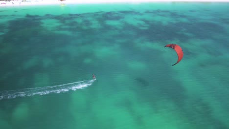 A-red-kite-surfer-glides-over-crystal-clear-waters-near-a-tropical-island,-aerial-view