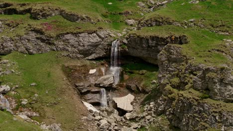 An-Einem-Sonnigen-Tag-Stürzt-Ein-Mehrstufiger-Bergwasserfall-Schroffe-Klippen-Und-Eisstücke-Hinab,-Touristen-Machen-Fotos,-Drohnen-Kamerafahrt