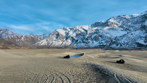 Dos-Vehículos-Todo-Terreno-Atraviesan-Un-Vasto-Paisaje-Desértico-Con-Majestuosas-Montañas-Cubiertas-De-Nieve-Como-Telón-De-Fondo