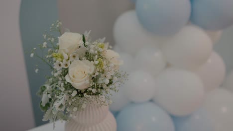 Zartes-Blumenarrangement-Aus-Weißen-Rosen-Und-Schleierkraut-In-Einer-Vase,-Vor-Einem-Hintergrund-Aus-Weißen-Und-Blauen-Luftballons